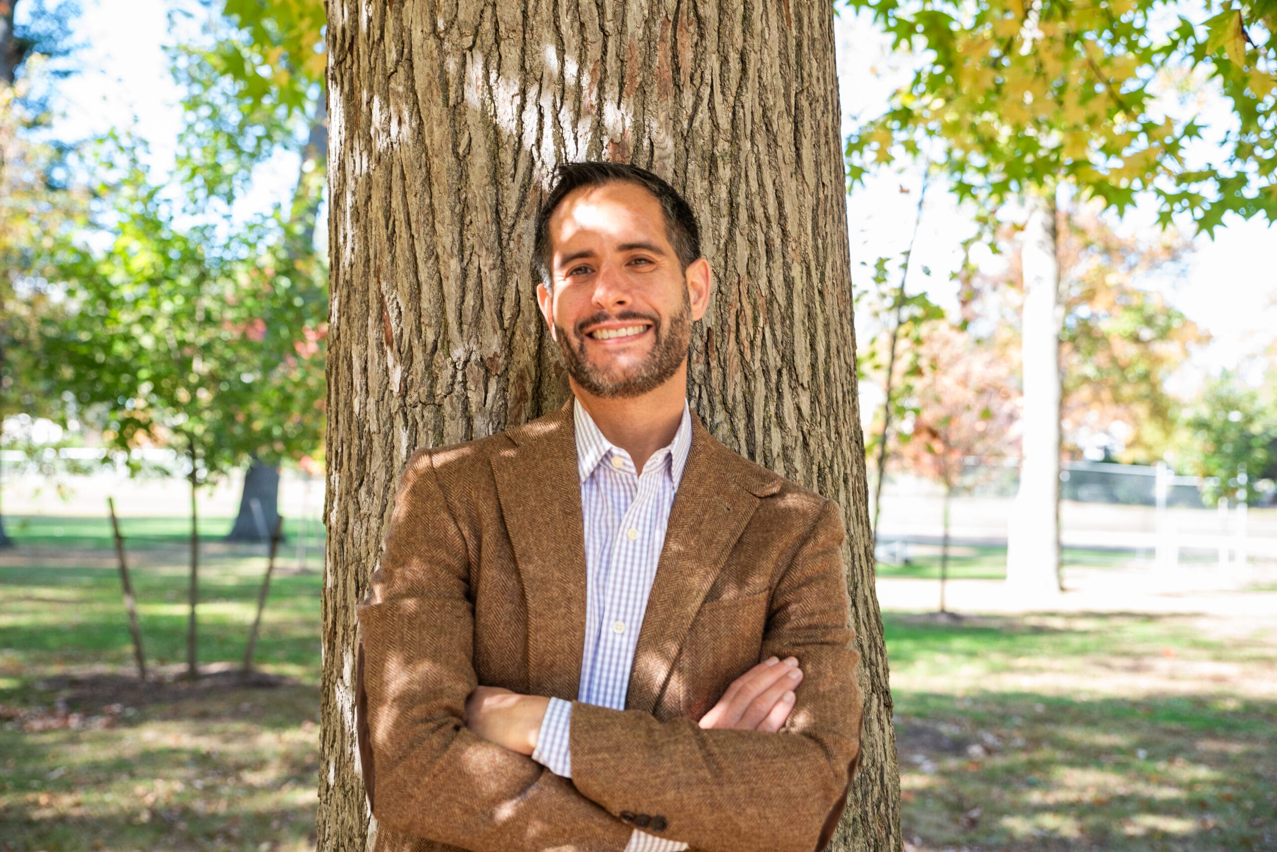 A man in a brown blazer smiles with arms crossed, leaning against a tree in a sunny park.
