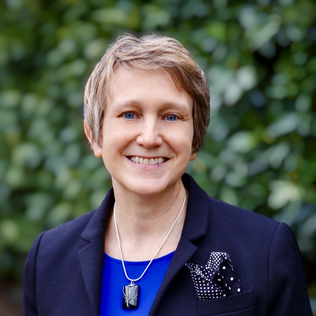 Smiling person with short hair in a black blazer and blue top, standing in front of green foliage.