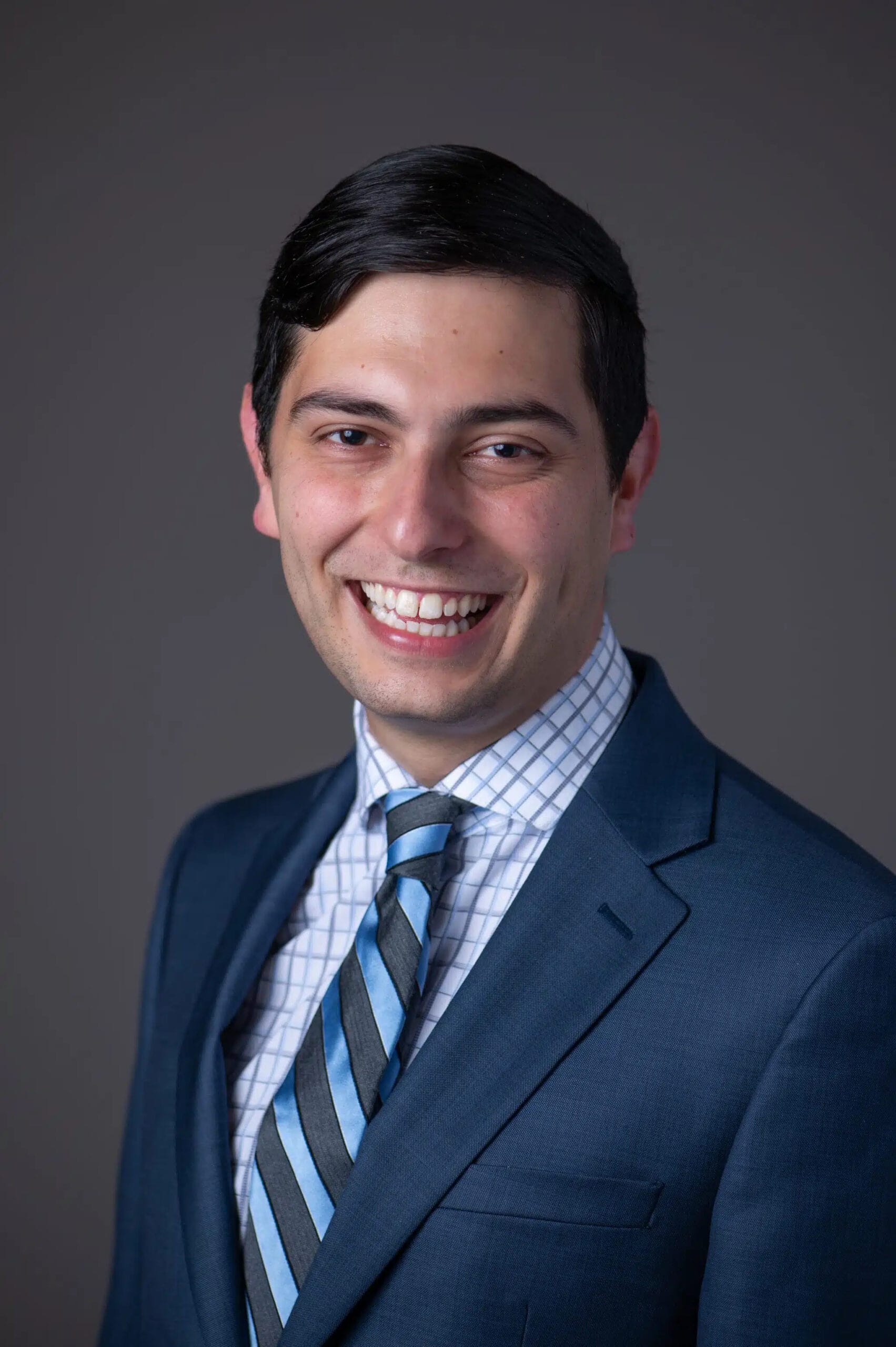 Smiling man in a blue suit, striped tie, and checkered shirt against a plain gray background.