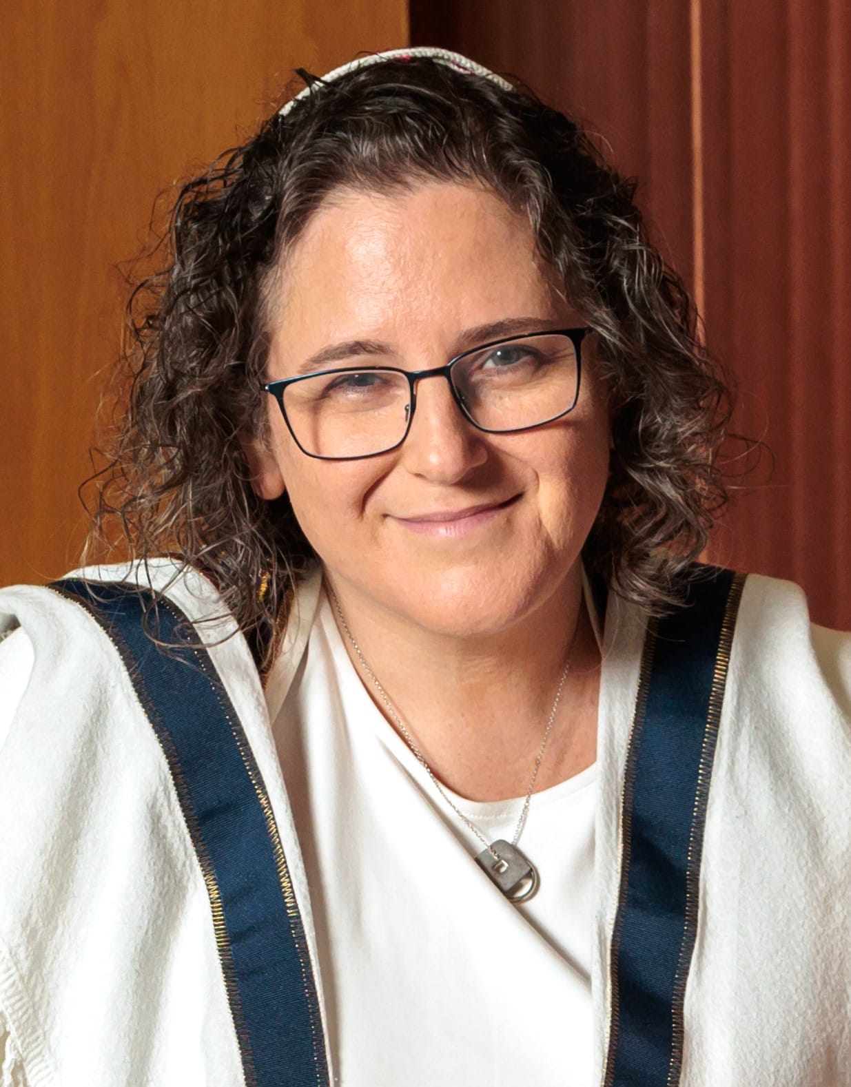Smiling person with curly hair, glasses, and a white robe with a blue stripe, standing indoors.
