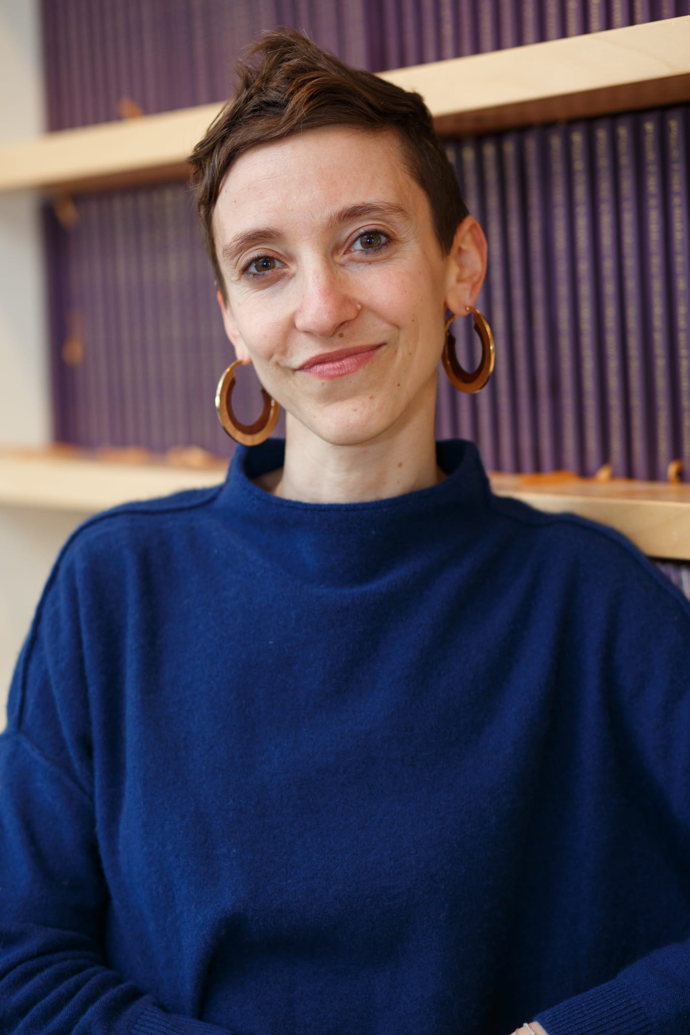 Person with short hair and hoop earrings smiling, wearing a blue sweater, standing by shelves of purple books.