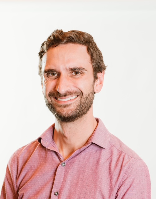 Man with short brown hair and beard, wearing a light pink collared shirt, smiling at the camera.