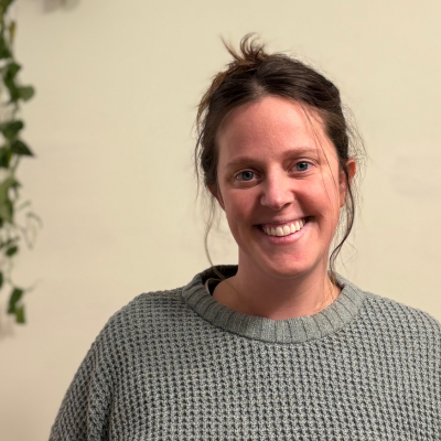 Woman with brown hair in a messy bun, smiling, wearing a grey sweater, standing indoors near a plant.