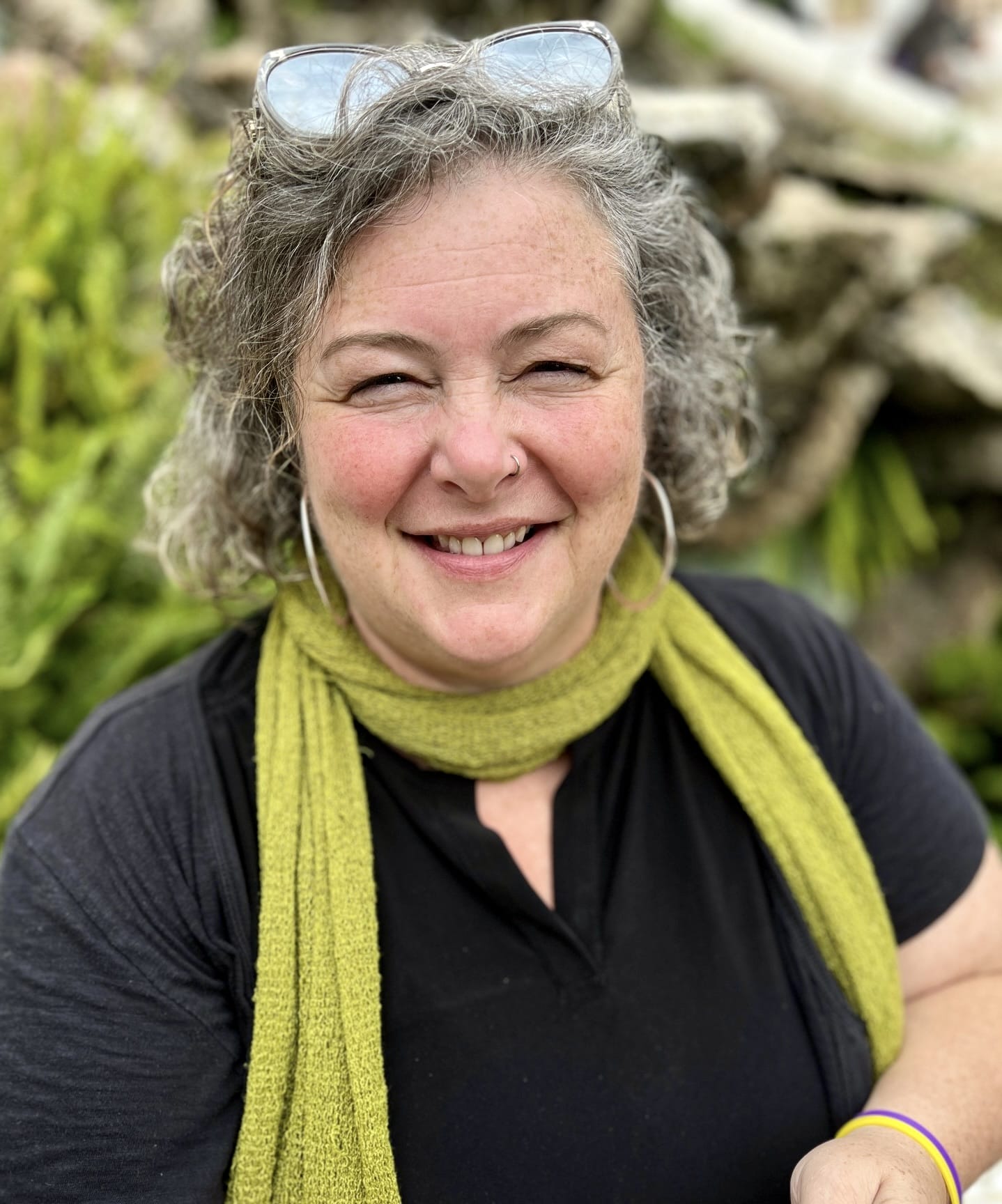Smiling woman with curly gray hair, black top, green scarf, and glasses on her head, outdoors.