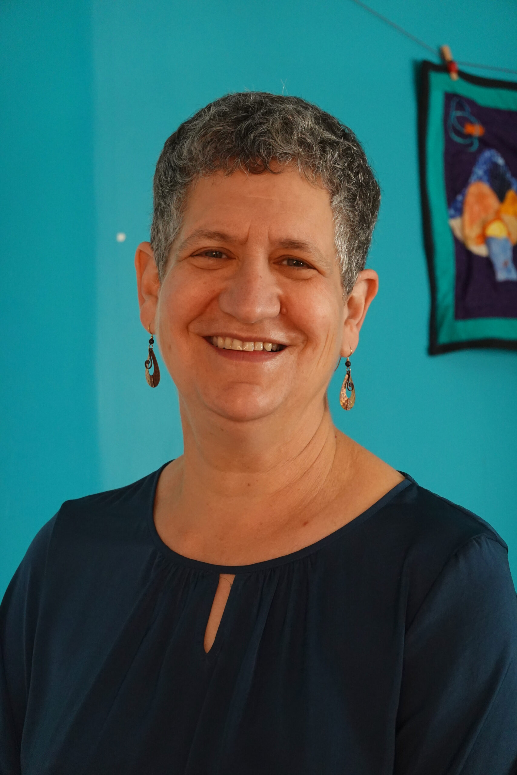 Smiling person with short curly gray hair and blue shirt against a teal wall with colorful art in the background.