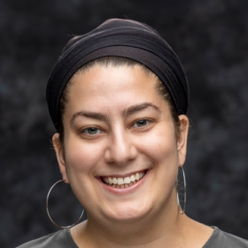 Woman smiling, wearing a black headscarf, large hoop earrings, and a dark top, with a dark background.