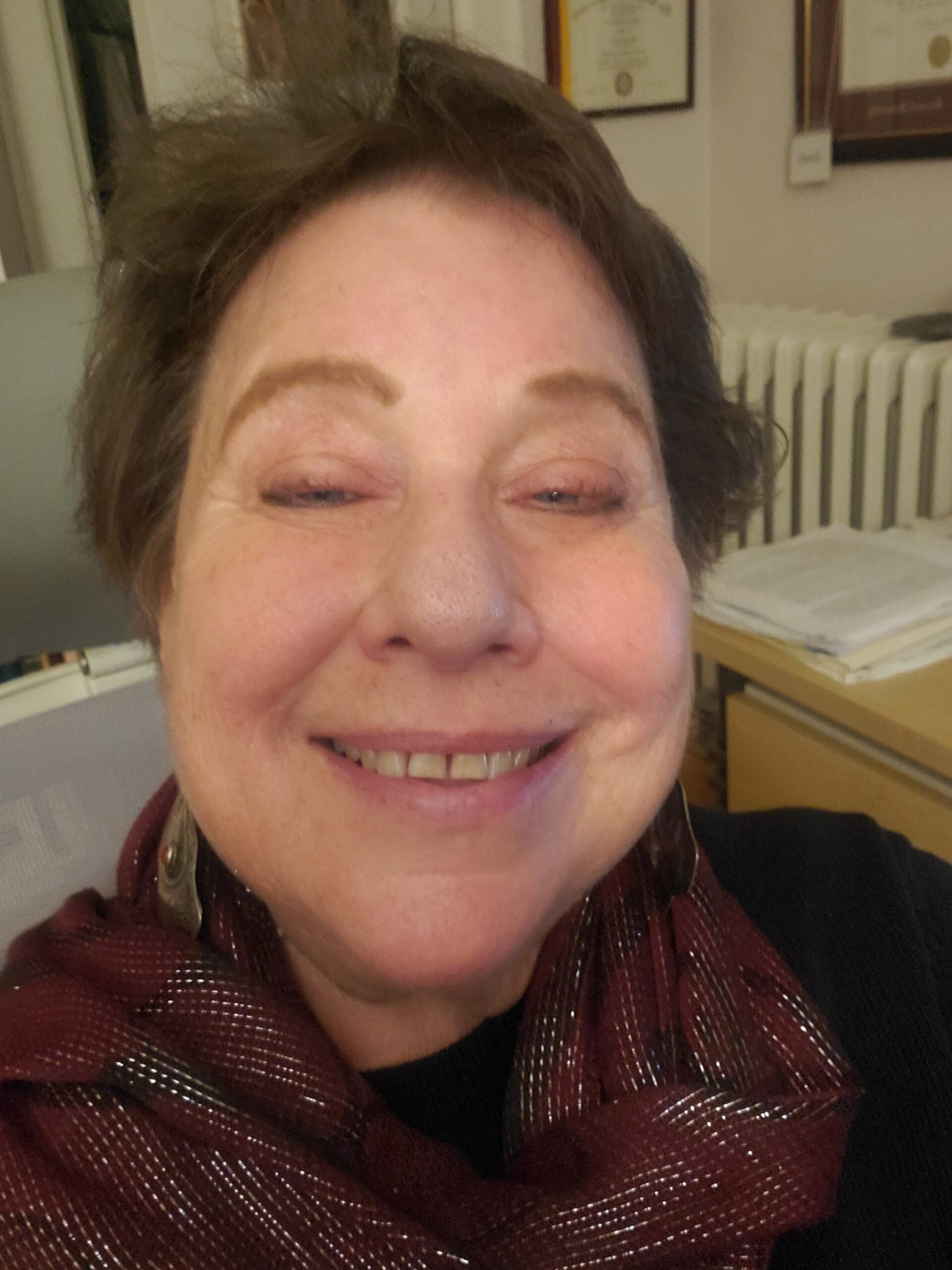 A woman with short brown hair smiles indoors, wearing a maroon scarf and earrings.