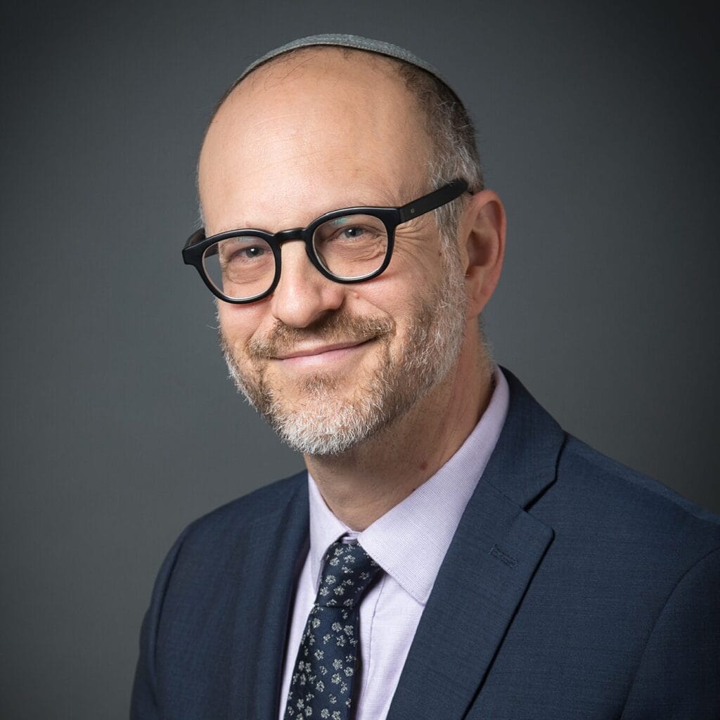 A smiling man with glasses and a beard, wearing a suit and tie, poses against a gray background.