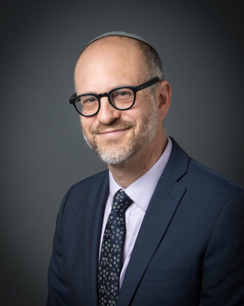 Bald man with glasses in a suit and tie, smiling in front of a dark gray background.