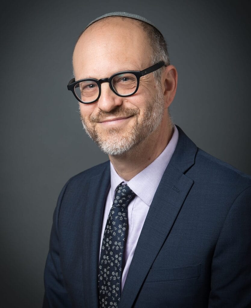 Bald man with glasses in a suit and tie, smiling in front of a dark gray background.