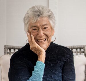 Smiling elderly woman with gray hair, wearing a dark top, rests her face on her hand, sitting on a chair.