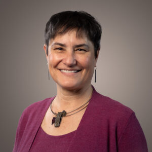 A smiling woman with short dark hair wearing a plum-colored top and a pendant necklace.