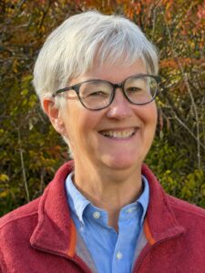 Smiling older woman with gray hair and glasses wearing a red jacket, outdoors with autumn foliage behind her.