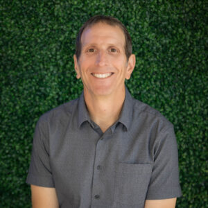 Smiling man in a short-sleeved shirt sits in front of a green leafy background.